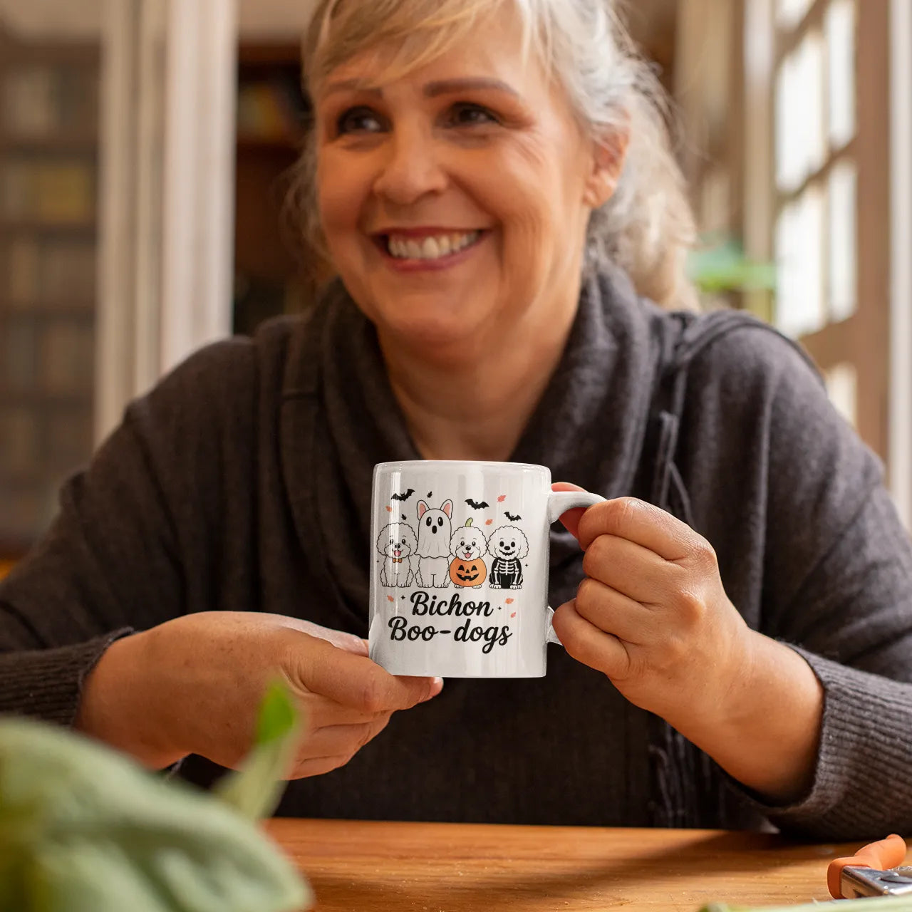 A happy woman holding a white Halloween-themed mug with cute Bichon Frise dogs in ghost and pumpkin costumes, sitting at a cozy wooden kitchen table.