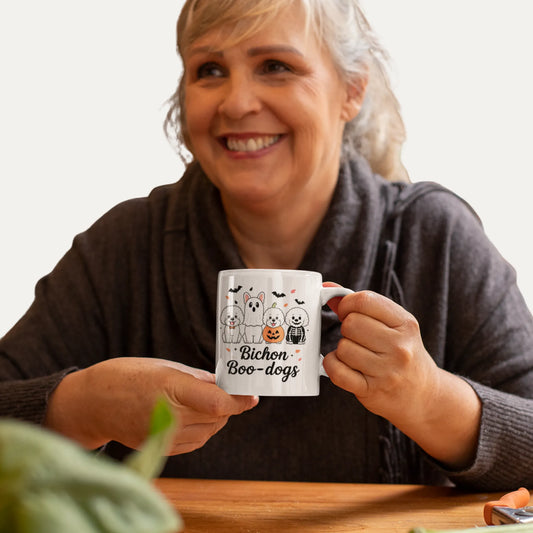 A happy woman holding a white Halloween-themed mug with cute Bichon Frise dogs in ghost and pumpkin costumes, sitting at a cozy wooden kitchen table.