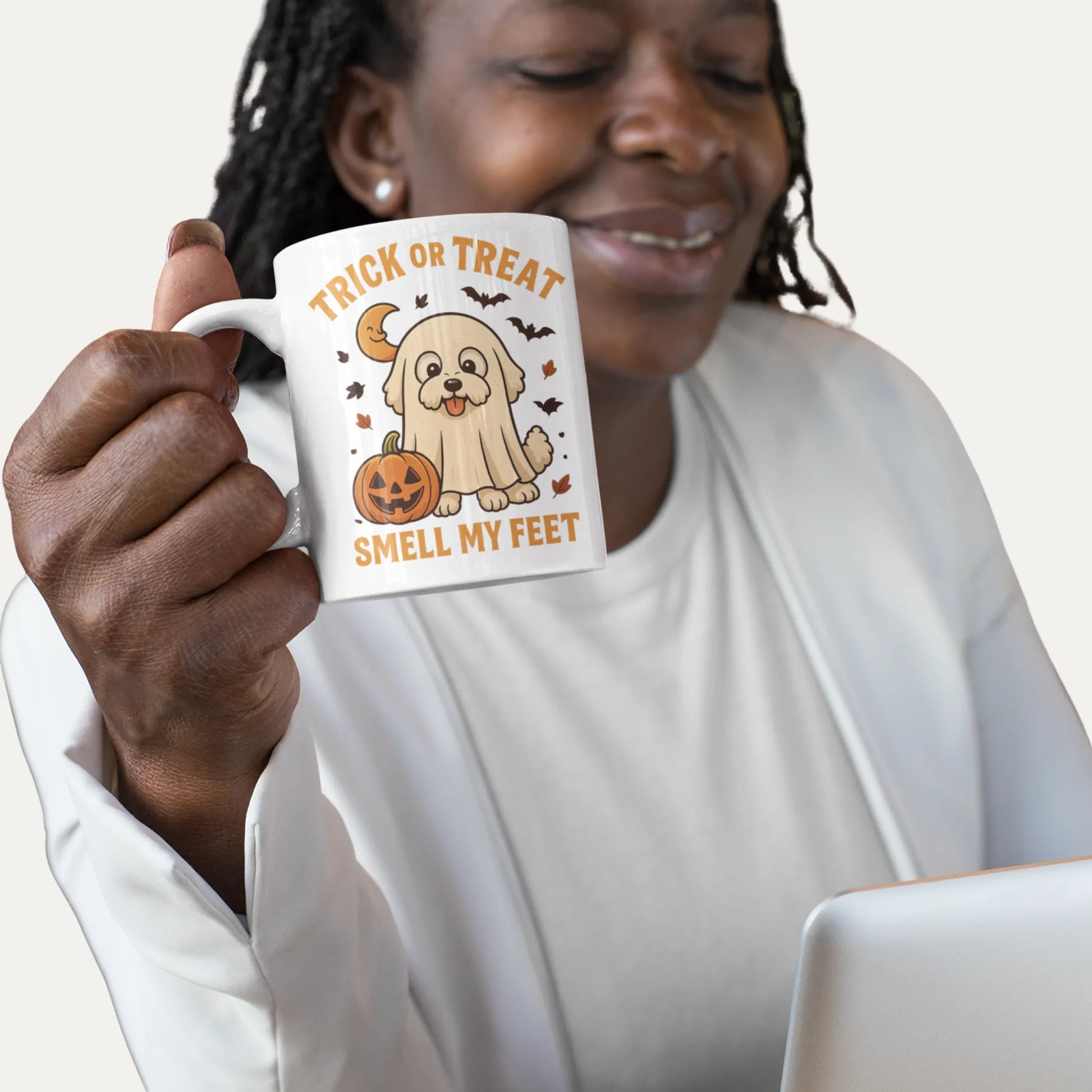 Smiling Black woman holding a white Halloween coffee mug with a Bichon Frise in ghost costume and pumpkin illustration, Trick or Treat Smell My Feet design