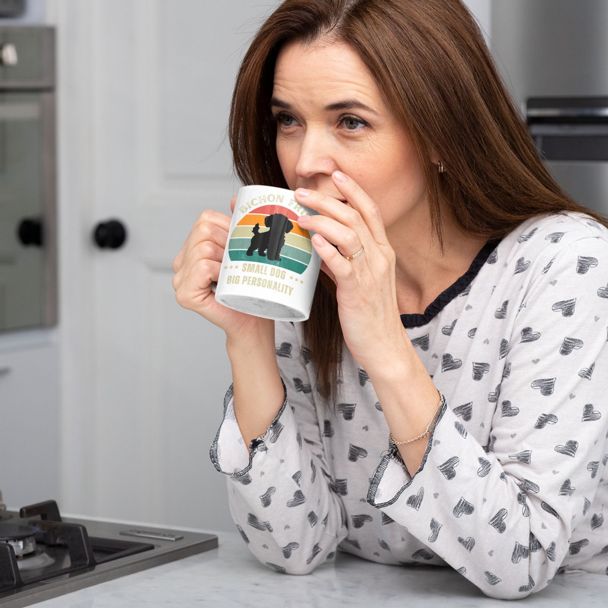 Middle-aged woman drinking from a white ceramic mug with retro Bichon Frise design and the text Small Dog Big Personality in a cozy kitchen setting