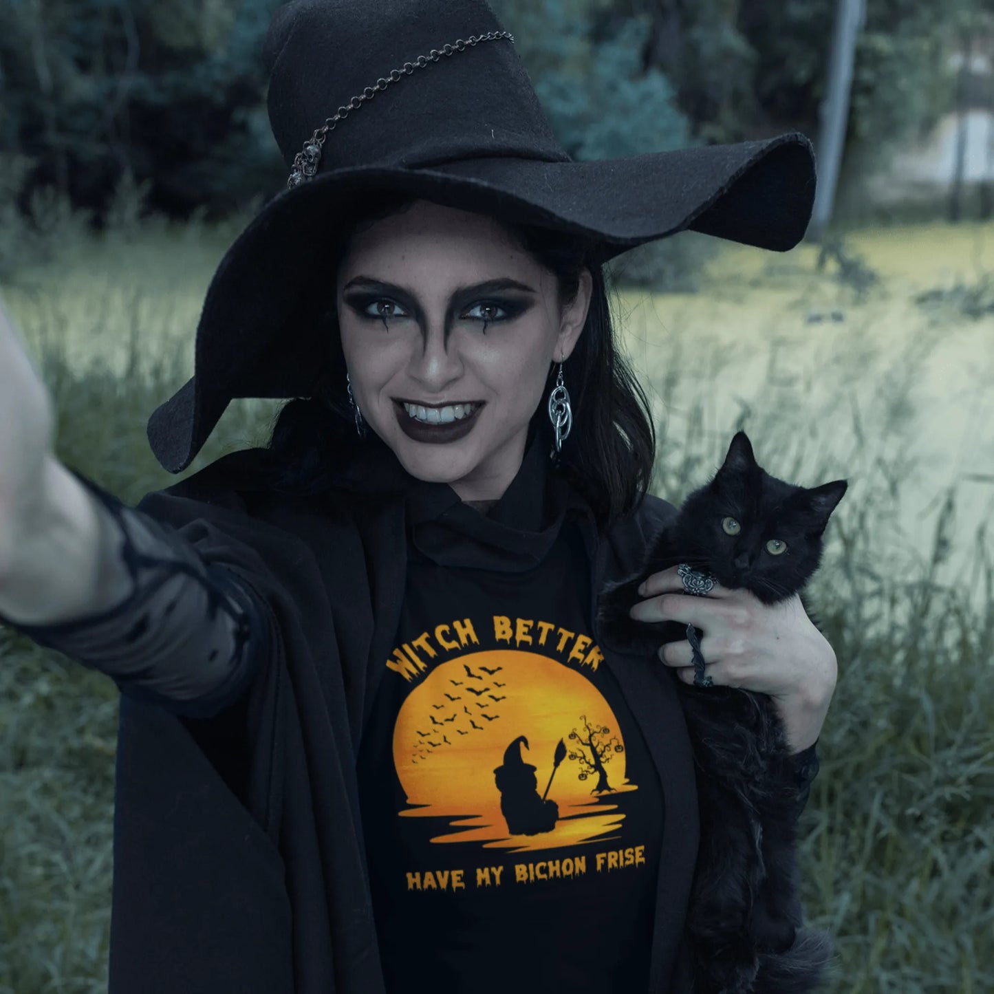 Woman in a witch costume holding a black cat and wearing a Halloween t-shirt that says “Witch Better Have My Bichon Frise” with a spooky sunset scene – perfect funny gift for Bichon Frise lovers.
